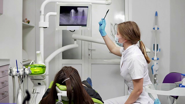一位年轻的女牙医正在椅子上的平板上展示x光的牙齿。女牙医戴着面具，穿着实验服。牙齿健康和牙齿保健。视频素材