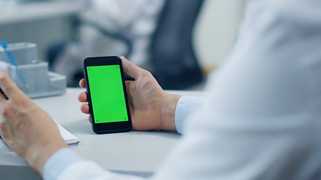 一个男性医生使用绿色屏幕的智能手机，在他的记事本上做笔记的特写。他的助手在他的桌子后面工作。办公室是白色和现代。视频素材