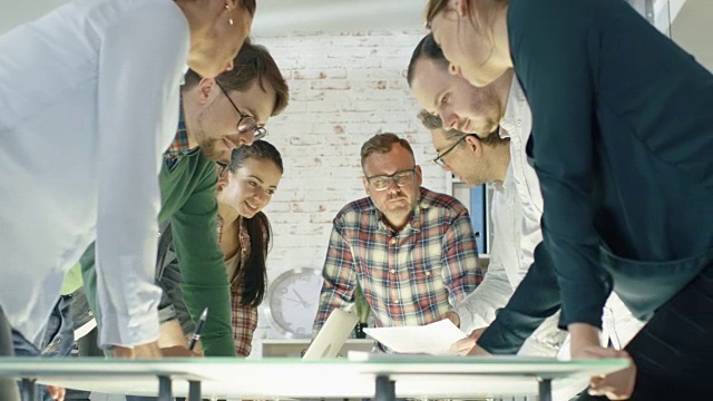 在创意办公室，富有成效的同事们站在桌子周围进行战略计划会议。他们在笔记本电脑上工作，交换笔记和想法。视频素材