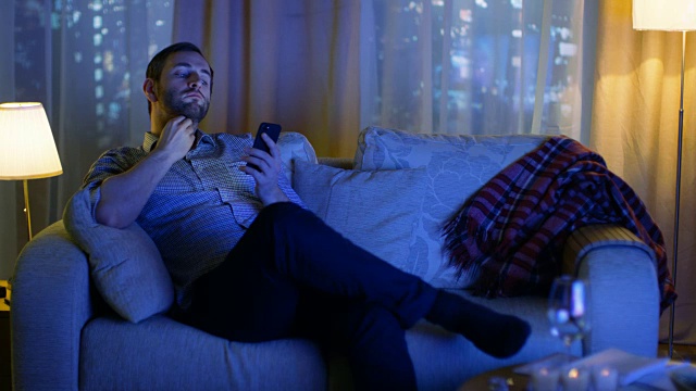 傍晚，中年男子坐在沙发上使用智能手机。打开电视照亮他。在一个有摩天大楼的大城市的背景窗口。视频素材