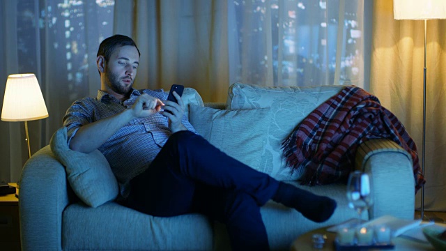 傍晚，中年男子坐在沙发上使用智能手机。打开电视闪光灯，照亮他。在一个有摩天大楼的大城市的背景窗口。视频素材