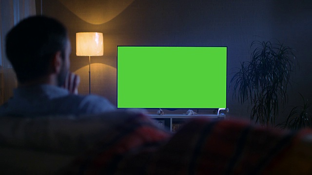 一个中年男子坐在沙发上看大屏幕电视的傍晚背影。视频素材