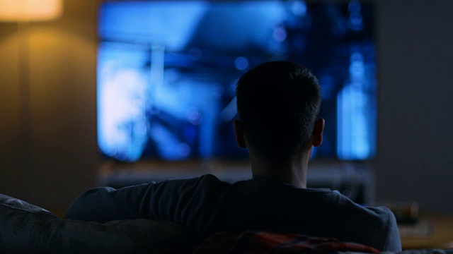 一个男人坐在沙发上看电影在他的大平板电视。视频素材