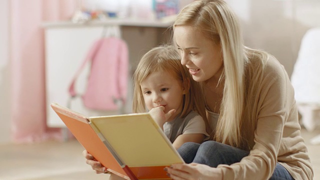 美丽的年轻妈妈和她可爱的小女儿一起读儿童读物。儿童的房间是粉红色的和充满了玩具。视频素材