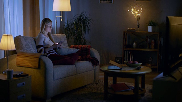 在晚上美丽的年轻女人放松在沙发上在她舒适的客厅。她使用智能手机。视频素材