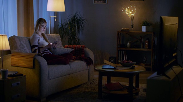 在晚上美丽的年轻女人放松在沙发上在她舒适的客厅。她一边用智能手机一边看电视。视频素材