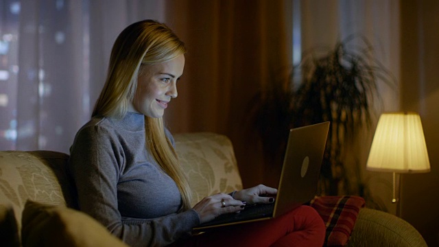 美丽的年轻女子在客厅用笔记本电脑工作，她坐在沙发上，膝盖上放着笔记本电脑。从窗口可以看到大城市。视频素材