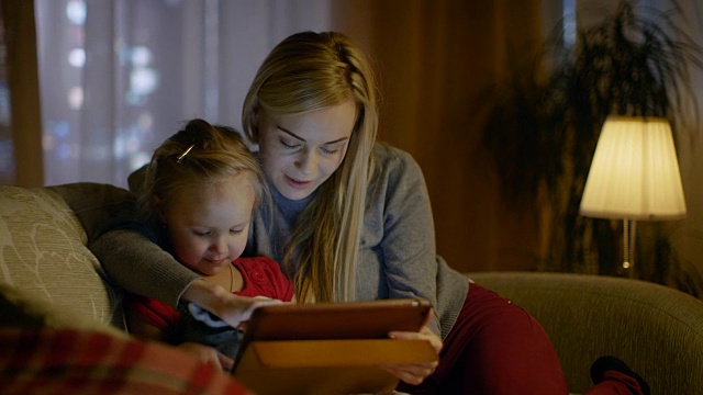 美丽的妈妈和她的小女儿正坐在客厅的沙发上，他们在用平板电脑。这是晚上，房间是舒适和温暖的。视频素材