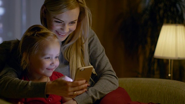 妈妈和她可爱的女儿正坐在客厅的沙发上。他们在使用智能手机和微笑。这是晚上。视频素材