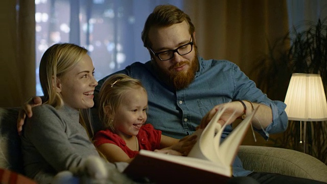爸爸、妈妈和小女儿在客厅的沙发上读儿童读物。这是晚上。视频素材