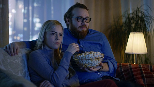 看电视。他们坐在舒适的客厅里的沙发上吃着爆米花。这是晚上。视频素材