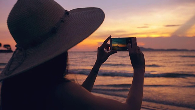 年轻女游客的剪影戴着帽子拍照与手机在日落在海洋海滩视频素材