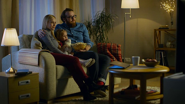 父亲，母亲和小女孩看电视的可能性不大。他们坐在舒适的客厅里的沙发上吃着爆米花。这是晚上。视频素材