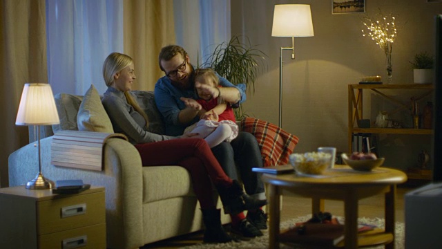 父亲，母亲和小女孩看电视的可能性不大。父亲向他的小女孩解释。他们坐在舒适的客厅里的沙发上。这是晚上。视频素材