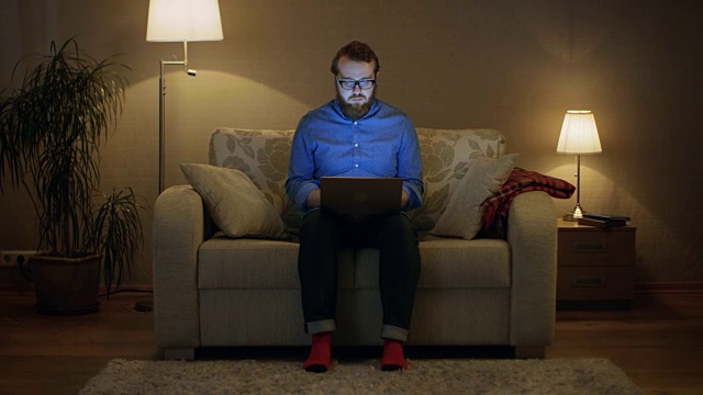 一个男人坐在客厅的沙发上，膝上放着笔记本电脑。他的工作。他直视着摄像机。落地灯打开。视频下载