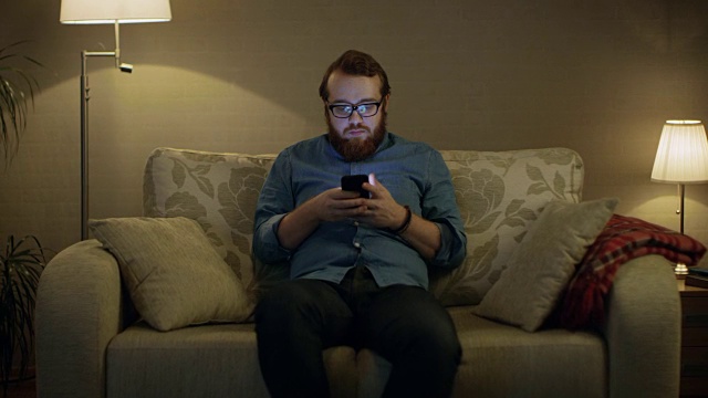 一个男人坐在客厅的沙发上，用智能手机拍照。落地灯打开。视频素材