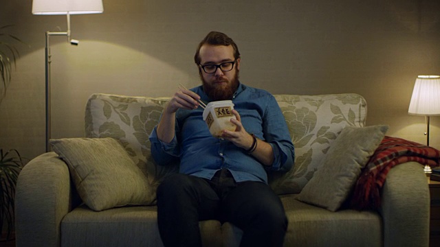一个男人坐在客厅的沙发上，从食物盒里吃亚洲食物。落地灯打开。视频素材