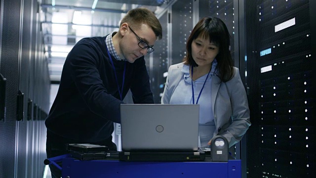 一名男性IT专家向一名女性服务器技术员展示笔记本电脑上的信息。他们正站在数据中心。视频素材