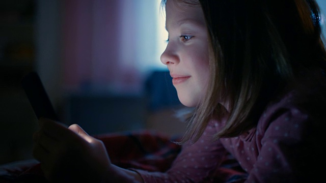 可爱的小女孩晚上在她的房间里，躺在床上使用智能手机。屏幕照亮她的脸。视频素材