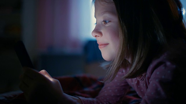 可爱的小女孩晚上在她的房间里，躺在床上使用智能手机。屏幕照亮她的脸。视频素材