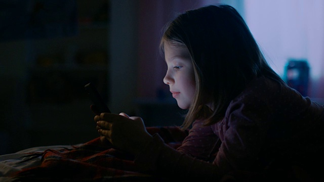 可爱的小女孩晚上在她的房间里，躺在床上使用智能手机。她的夜灯打开了。视频素材