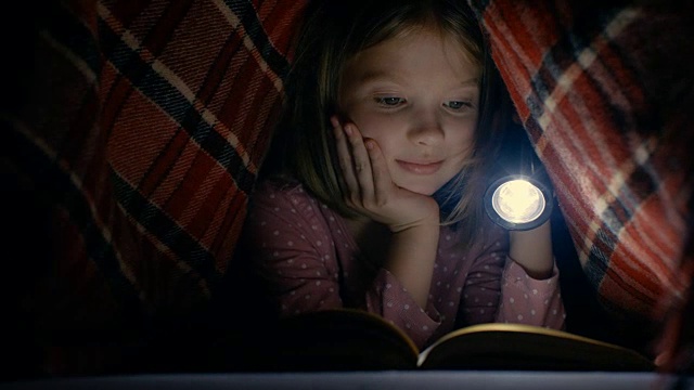 聪明的小女孩在毯子下用手电筒读有趣的书。视频下载