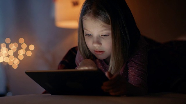 可爱的小女孩晚上躺在她的床上，她拿着平板电脑。她的夜灯开着。视频素材