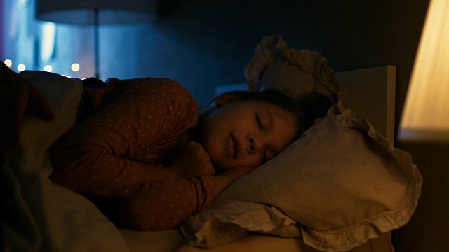 甜美的小女孩晚上睡在她的床上，她的妈妈盖好她的毯子。视频素材