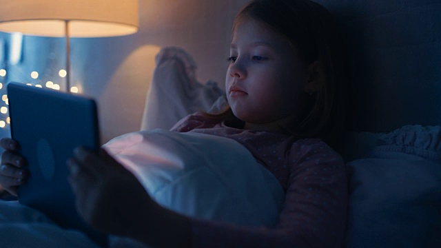 快乐的小女孩晚上躺在床上看平板电脑上的东西。她床上的灯是开着的。视频素材