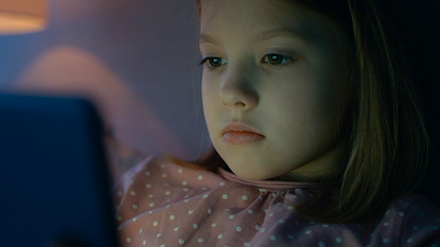 可爱的小女孩晚上躺在床上，用平板电脑照亮她的脸。视频素材