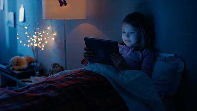 可爱的小女孩躺在床上用平板电脑。视频素材