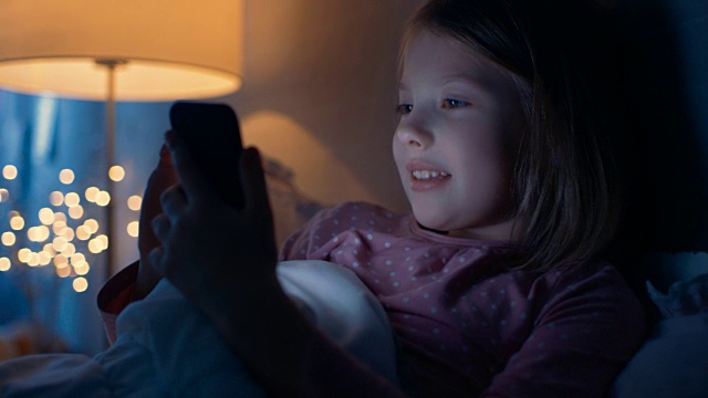 可爱的小女孩在床上与智能手机互动。视频素材
