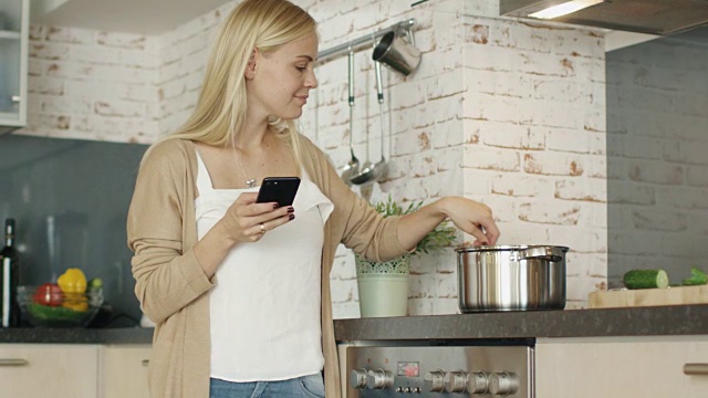 愉快的女人站在厨房灶台前使用智能手机。她在锅里搅拌着什么东西。视频素材