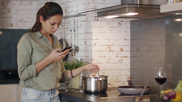 年轻女子一边拿着手机一边微笑着在平底锅里搅拌食物。视频素材