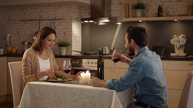 一对美丽的夫妇在厨房里吃烛光晚餐。他们喝酒、吃饭、聊天。微笑和心情都很好。视频素材