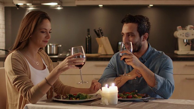 一对美丽的夫妇在厨房里吃烛光晚餐。他们吃喝聊天。庆祝时他们碰杯。视频素材