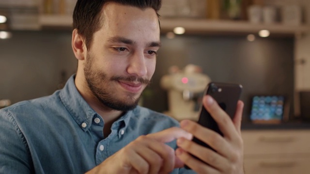 英俊的年轻人微笑着在厨房使用智能手机。视频素材