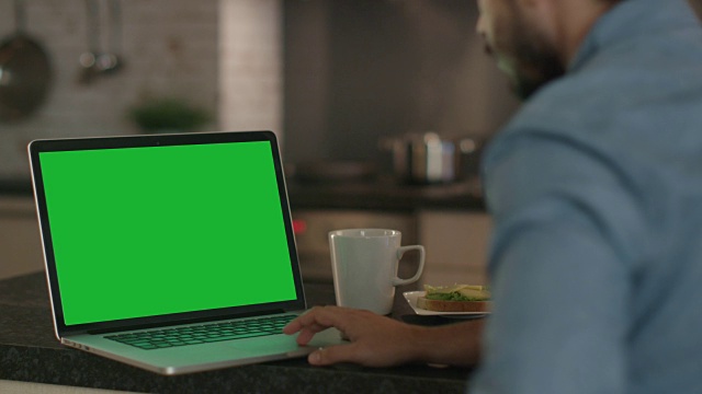 一位年轻人坐在餐桌旁使用绿色屏幕的笔记本电脑。视频素材