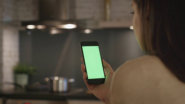 年轻女孩坐在厨房里使用绿屏智能手机。视频素材