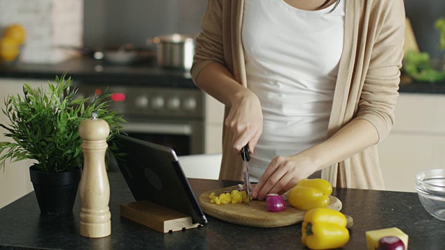 一个女人在木板上切蔬菜的特写。在平板电脑上查找食谱。视频素材