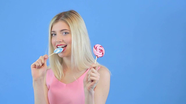 年轻女子舔着颜色发亮的棒棒糖，映衬着蓝色的背景视频素材