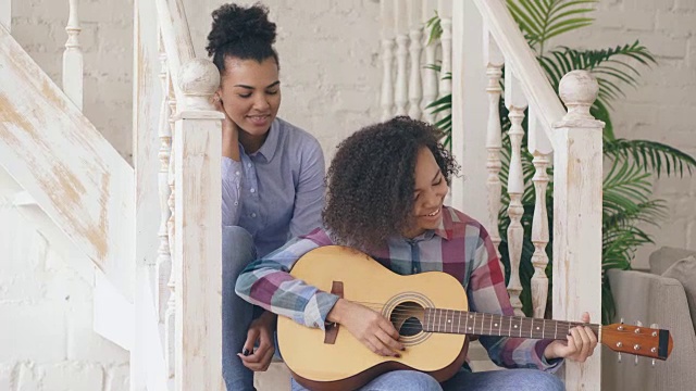 两个巴西卷发女孩坐在楼梯上练习弹原声吉他。朋友们玩得开心，在家里唱歌视频素材