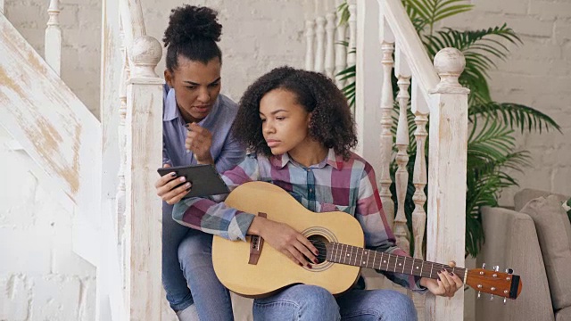 混血年轻女子坐在楼梯上教她十几岁的妹妹在家里弹原声吉他视频素材