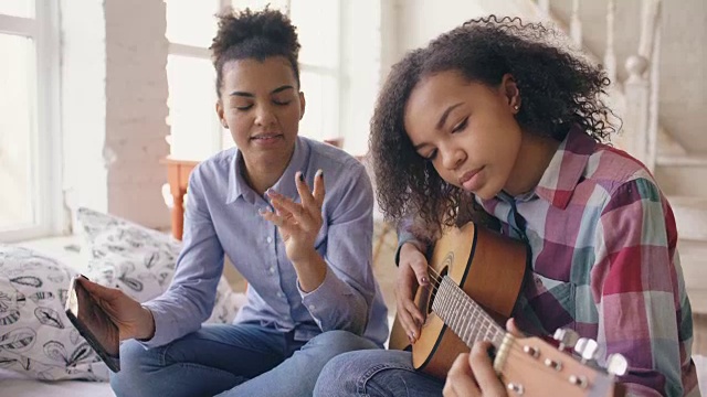 混合种族的年轻女子与平板电脑坐在床上教她十几岁的妹妹在家里弹吉他视频素材