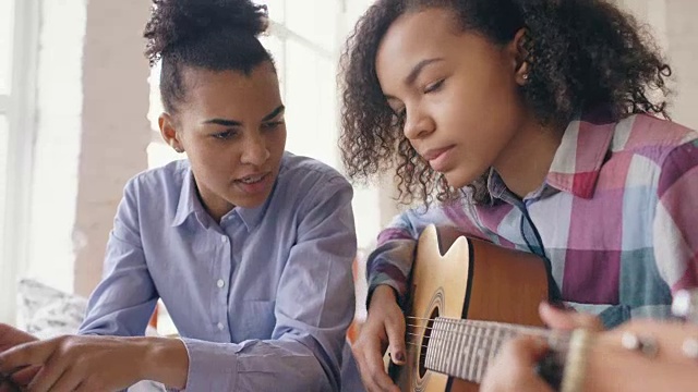 混合种族的年轻女子与平板电脑坐在床上教她十几岁的妹妹在家里弹吉他视频素材