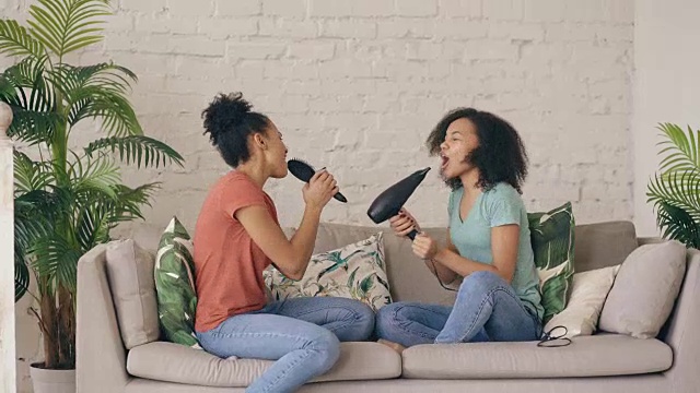 混合种族年轻有趣的女孩跳舞唱歌与吹风机和梳子坐在沙发上。姐妹们有乐趣休闲在客厅在家里的概念视频素材