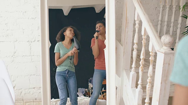 混合种族的年轻有趣的女孩跳舞唱歌与吹风机和梳子在镜子前。姐妹有乐趣休闲在卧室在家里的概念视频素材