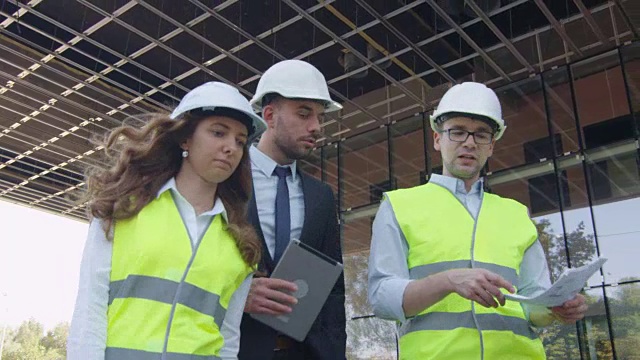 两个工程师和商人戴着安全帽走路，说话，使用平板电脑的团队。背景中的玻璃建筑或摩天大楼。视频素材