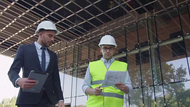 工程师和商人戴着安全帽走路、说话和使用平板电脑。背景中的玻璃建筑或摩天大楼。视频素材