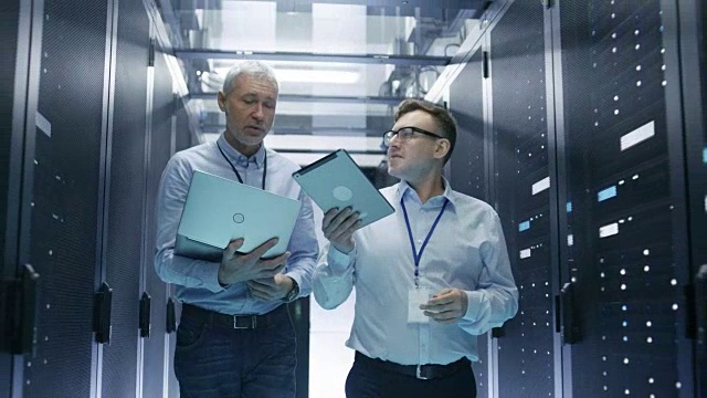 在数据中心，两个IT工程师穿过一排排的服务器机架。他们在平板电脑和笔记本电脑上工作。视频素材